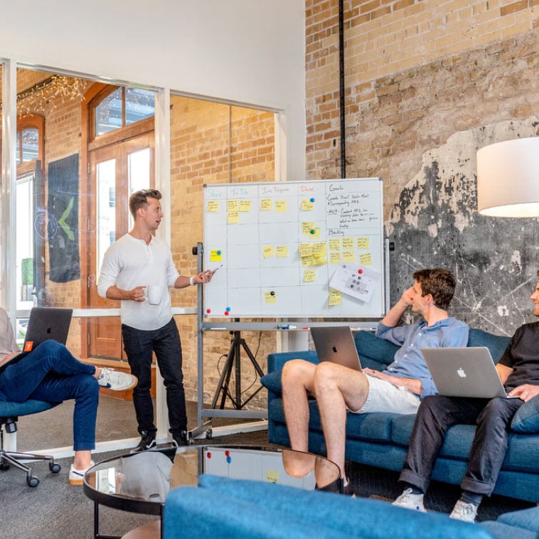 A diverse group of individuals sitting around a table with laptops, engaged in a meeting or collaboration session.