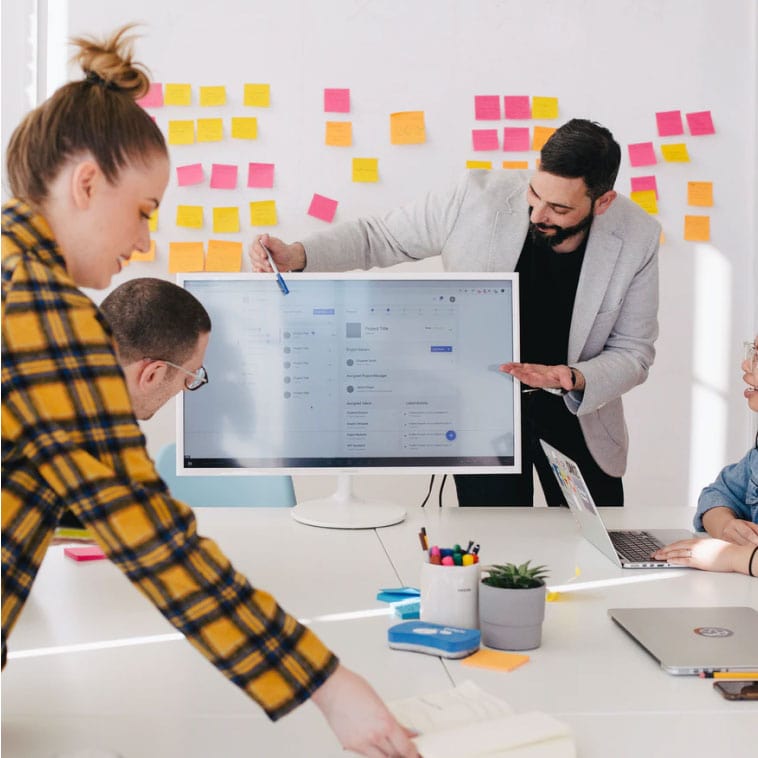 A diverse group of professionals in an office, gathered around a computer screen for a meeting.
